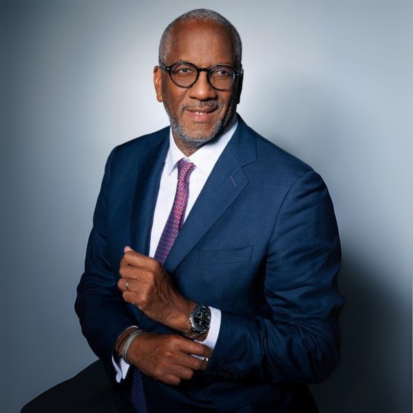 Professional headshot of a senior business executive wearing a navy blue suit, glasses, and a pink tie, smiling at the camera.